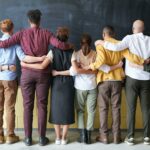 A diverse group of adults in casual outfits hugging in front of a chalkboard, symbolizing teamwork.
