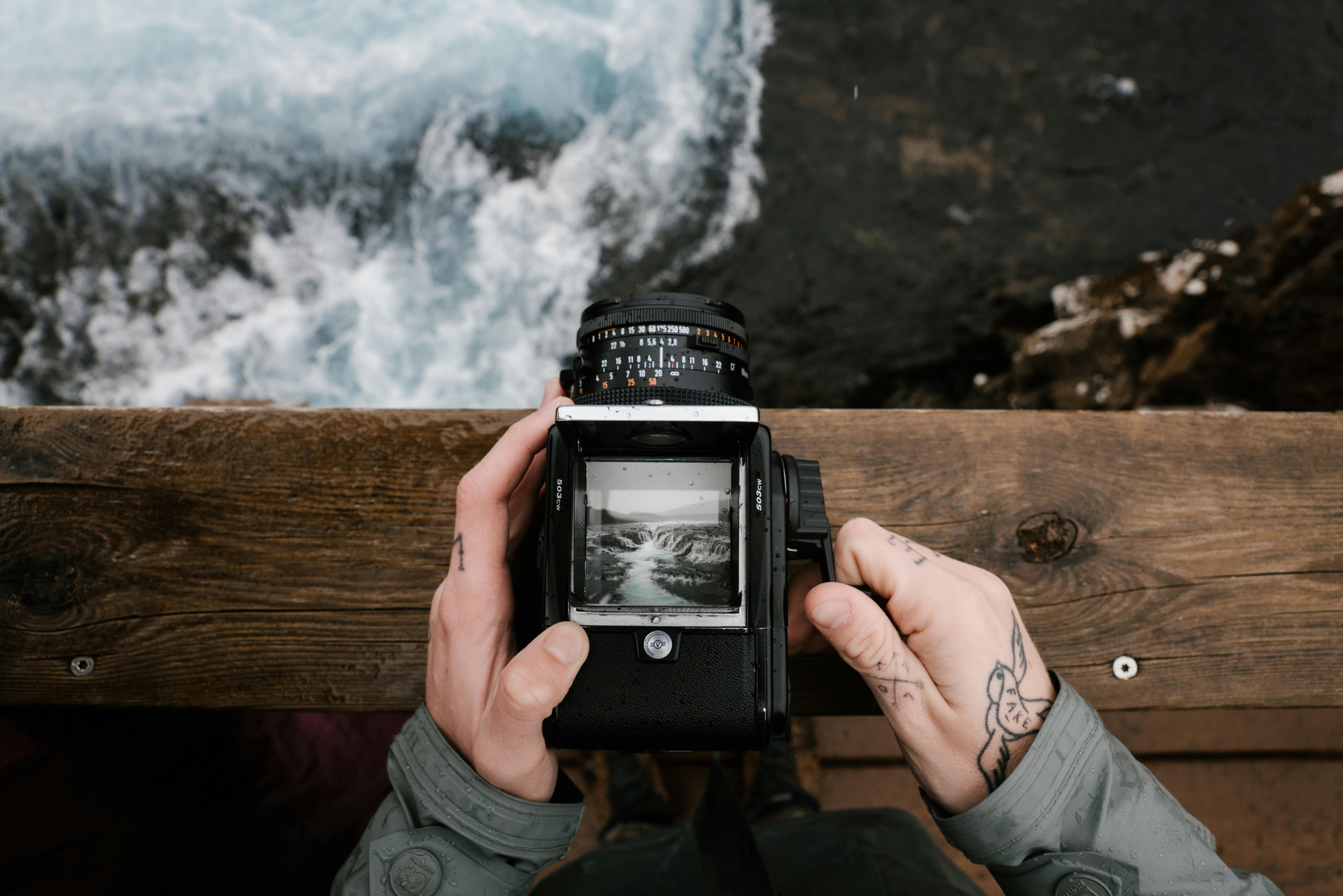 Close-up of hands holding a camera while standing on a wooden railing, overlooking rough waters below.