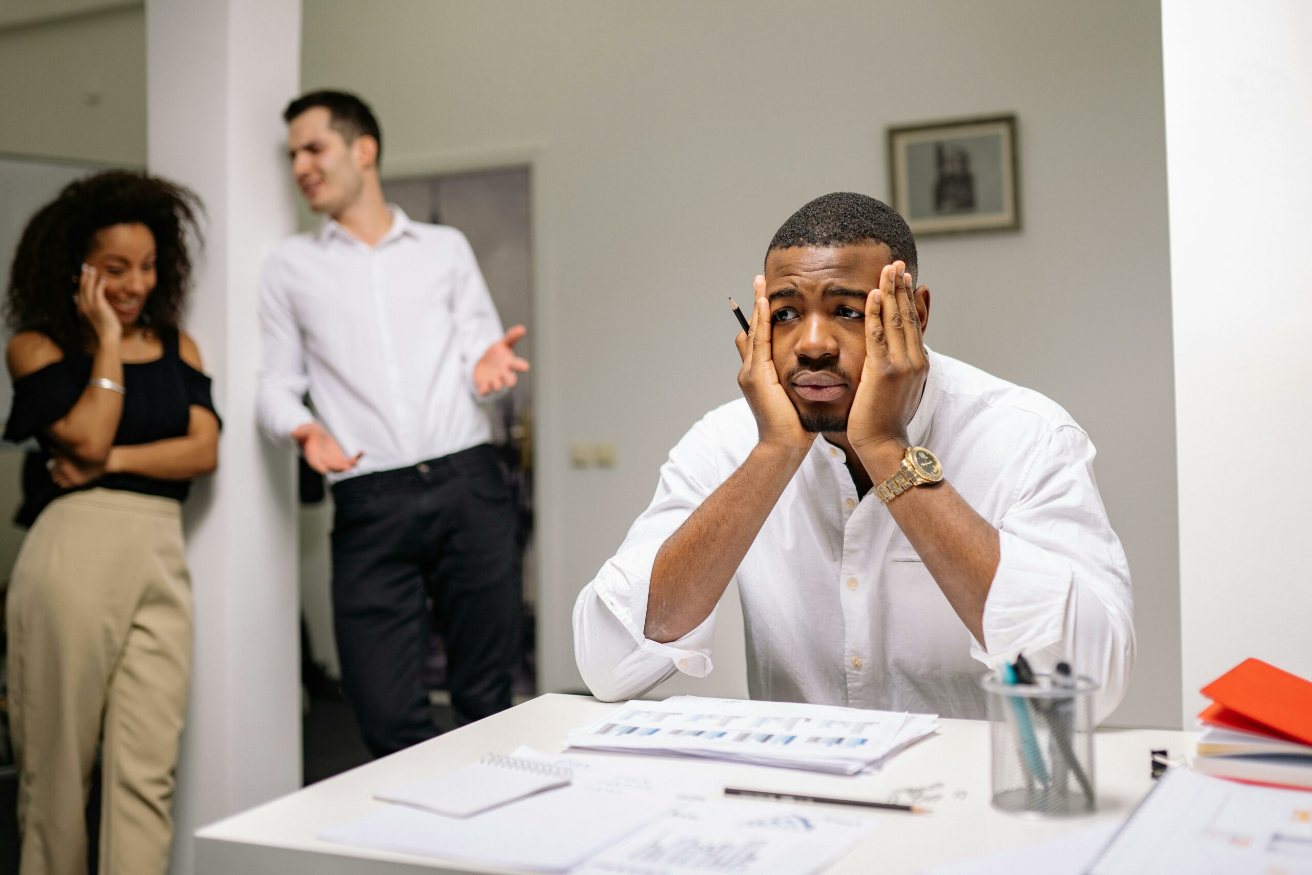 A stressed employee sits at a desk with colleagues in discussion nearby, highlighting workplace tension.